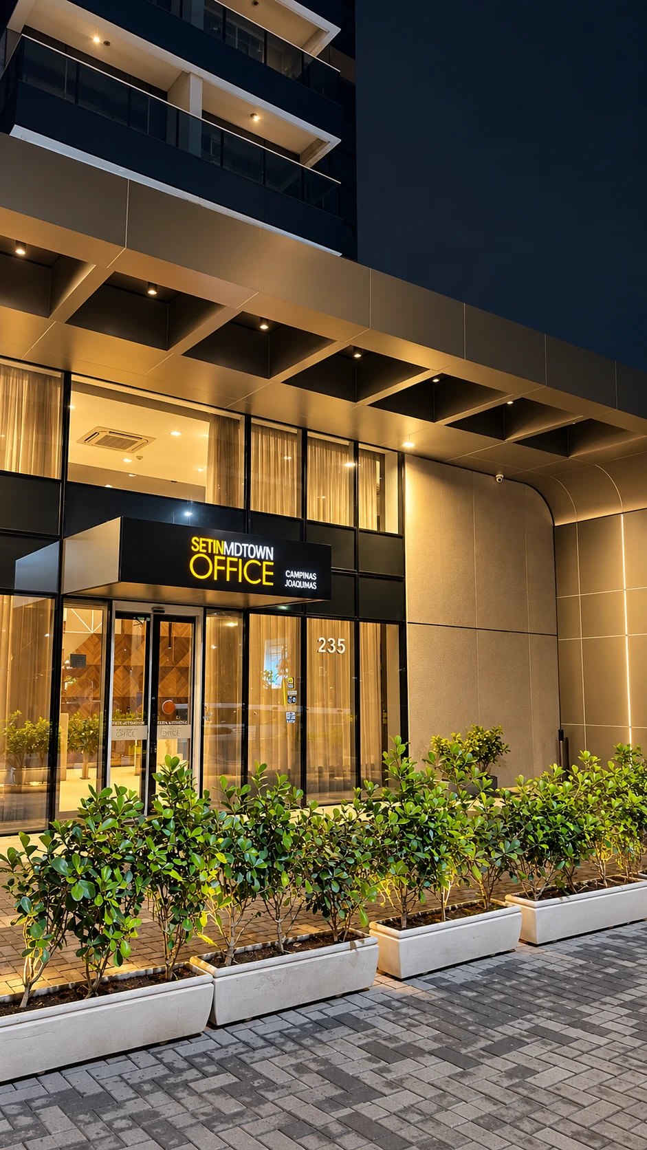 Fachada iluminada do Setin Midtown Office, edifício onde fica o consultório, com entrada de vidro e canteiros de plantas.